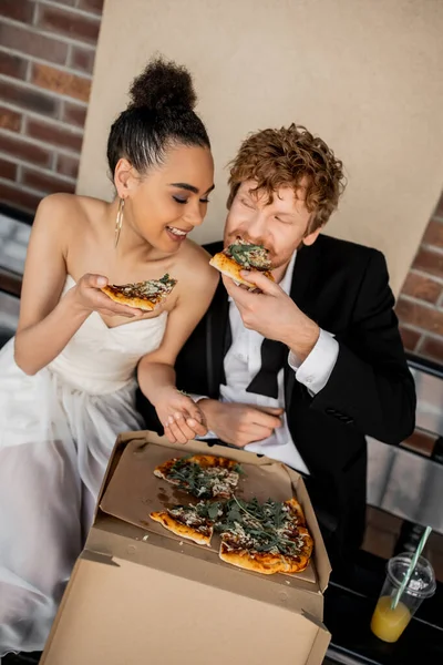 Pleased multiethnic couple in wedding attire eating tasty pizza on bench, wedding in city — Stock Photo