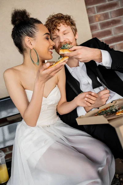 Стильная межрасовая пара в свадебном наряде ест вкусную пиццу на скамейке в городе, свадьба на открытом воздухе — стоковое фото