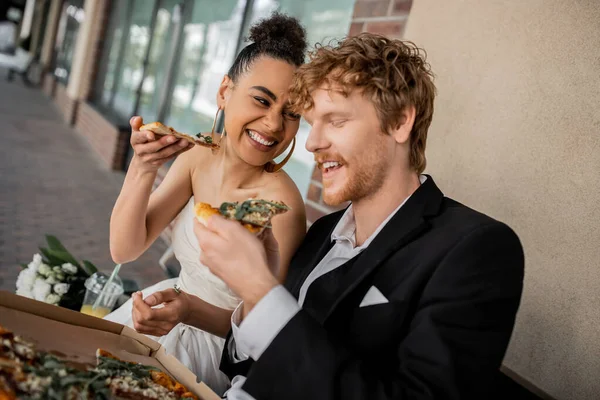 Весела афроамериканська наречена з піцою біля молодого рудого нареченого, весілля в місті — стокове фото