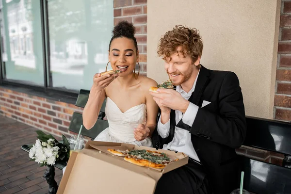 Smiling african american bride and redhead groom eating pizza on bench, wedding celebration in city — Stock Photo