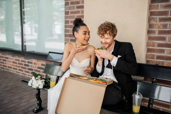 Cheerful, elegant multiethnic newlyweds eating pizza near flowers and orange juice on bench in city — Stock Photo
