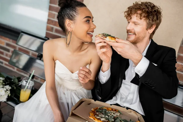 Boda al aire libre, diversión, pelirroja novio alimentación alegre afroamericana novia en el banco en la ciudad - foto de stock