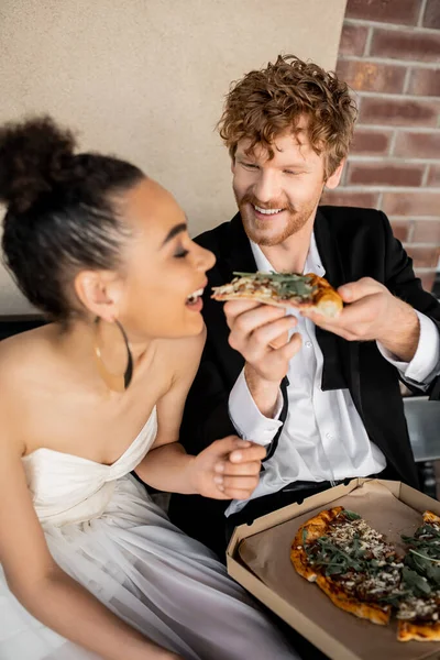 Redhead groom feeding overjoyed bride with tasty pizza on bench, outdoor wedding in european city — Stock Photo