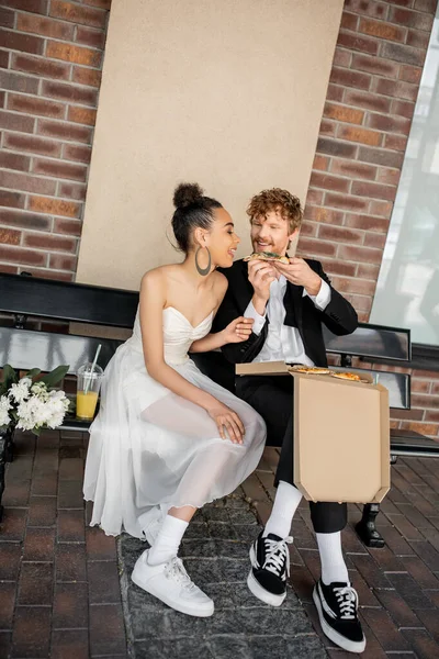 Stylish and happy interracial newlyweds eating pizza near orange juice and flowers on bench in city — Stock Photo