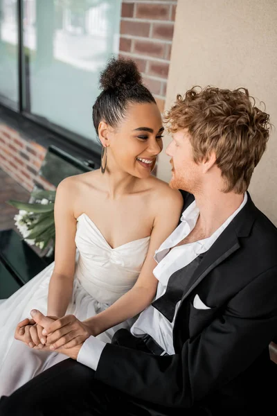 Cheerful interracial couple holding hands and looking at each other on bench, wedding in city — Stock Photo