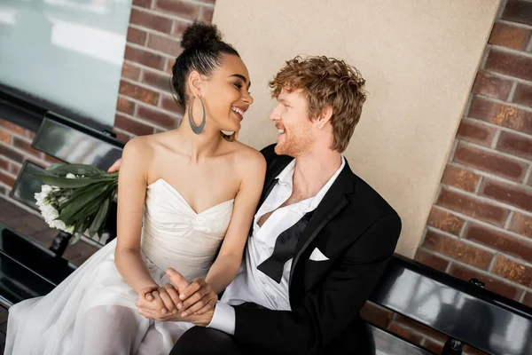 Joven pareja multiétnica sonriendo el uno al otro y tomados de la mano en el banco, boda, entorno urbano - foto de stock