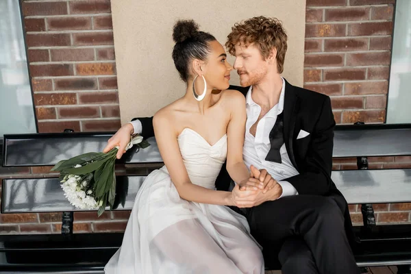 Pelirrojo con flores sentado cara a cara con la novia afroamericana en el banco, boda al aire libre - foto de stock