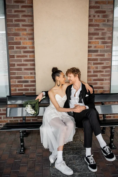 Mariage en ville, heureux marié rousse avec bouquet assis face à face avec la mariée afro-américaine — Photo de stock