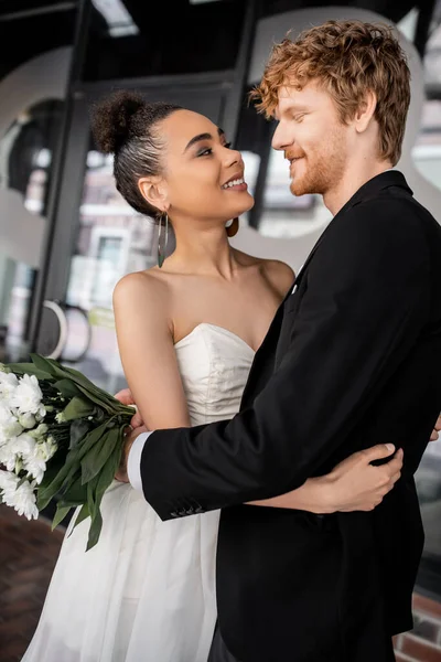 Super alegre e elegante casal interracial em vestido de casamento abraçando na rua da cidade — Fotografia de Stock