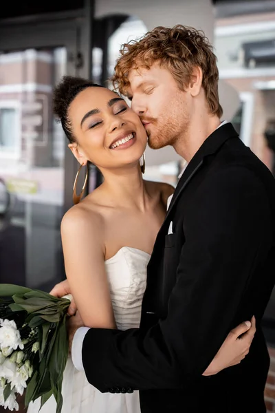 Redhead groom in black suit embracing and kissing stylish african american bride on city street — Stock Photo