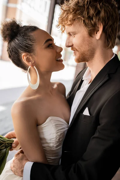Joyful and elegant interracial newlyweds looking at each other on street, wedding in urban setting — Stock Photo