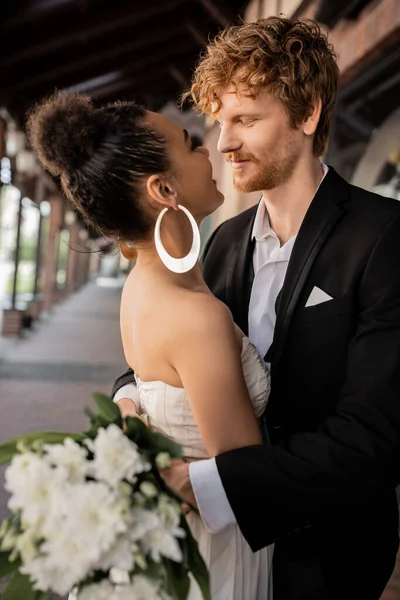 Gioiosa coppia interrazziale che si abbraccia e si guarda, abito da sposa, ambiente urbano — Foto stock