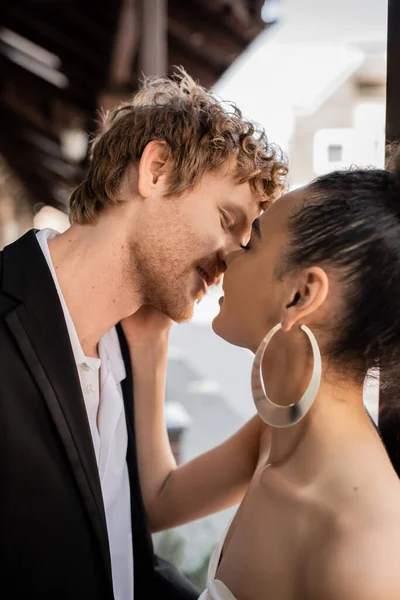 Novia afroamericana besando novio pelirrojo en traje negro, vista lateral, boda en la ciudad - foto de stock