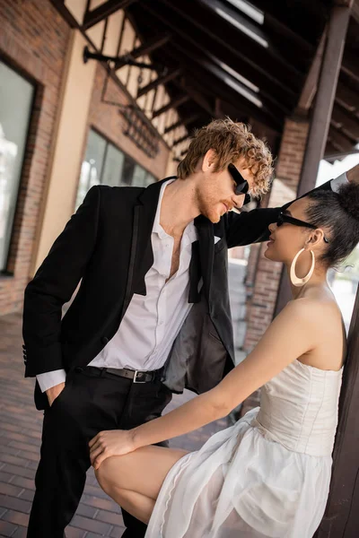 Стильний наречений в сонцезахисних окулярах, з рукою в кишені біля елегантної афроамериканської нареченої на міській вулиці — стокове фото