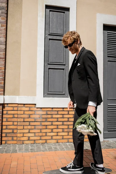 Елегантний чоловік в чорному костюмі і сонцезахисних окулярах, що йдуть по міській вулиці, наречений з весільним букетом — стокове фото