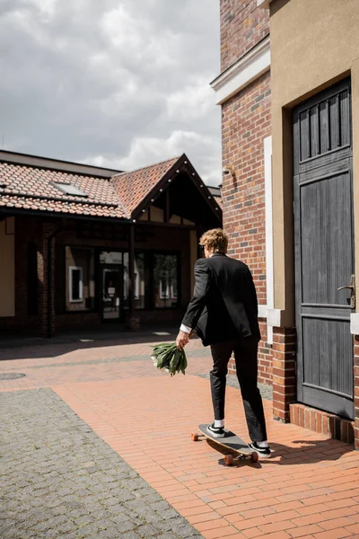 Back view of groom in black suit holding wedding bouquet and  riding longboard on city street — Stock Photo