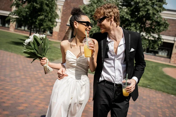 Joyful multiethnic newlyweds in sunglasses walking with orange juice, wedding on urban street — Stock Photo