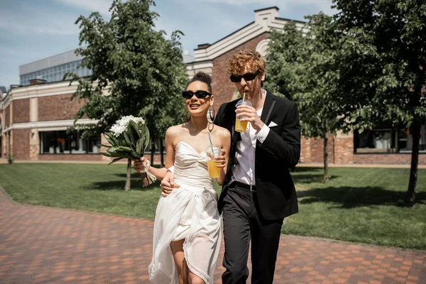 Elegant newlyweds in sunglasses walking with orange juice and flowers in european city — Stock Photo
