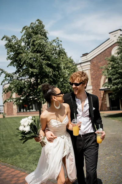 Разнообразная пара с апельсиновым соком и цветами, улыбающиеся друг другу, свадьба на улице — стоковое фото
