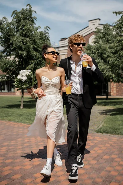 Happy interracial couple, wedding attire, sunglasses, orange juice, bouquet, wedding in city — Stock Photo