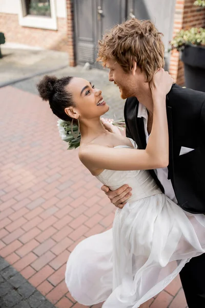 Super alegre e elegante casal interracial em trajes de casamento abraçando na rua urbana — Fotografia de Stock