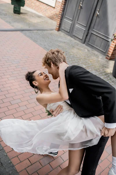 Elegante pelirroja hombre abrazando alegre africana americana novia, boda en la calle urbana - foto de stock
