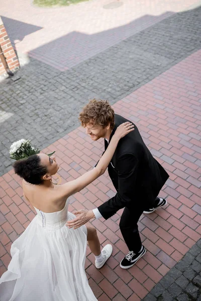 Feliz casal interracial em traje de casamento correndo para o outro na rua urbana, vista superior — Fotografia de Stock