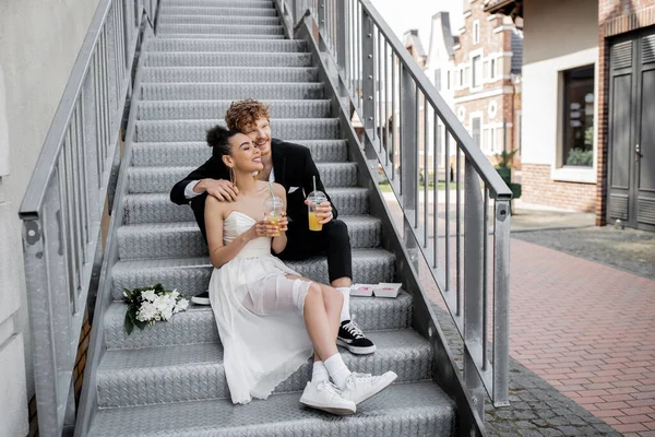 Multiethnic newlyweds with orange juice sitting on stairs and looking away, wedding in city — Stock Photo