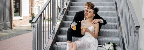 Hochzeit auf der Straße, zufriedenes gemischtes Paar sitzt mit Orangensaft auf der Treppe, Transparent — Stockfoto