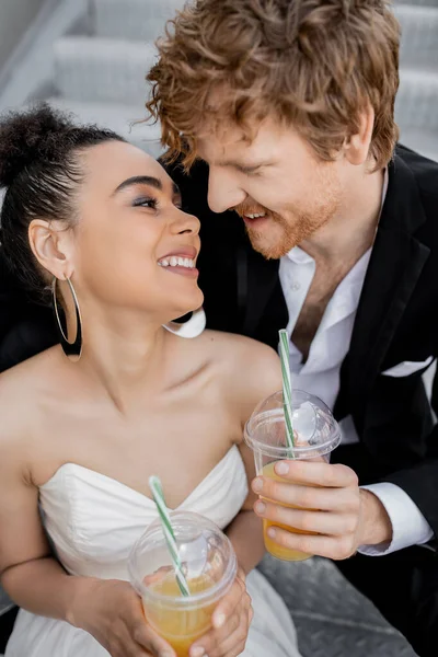 Coppia interrazziale felice in abito da sposa, con succo d'arancia, guardarsi all'aperto — Foto stock