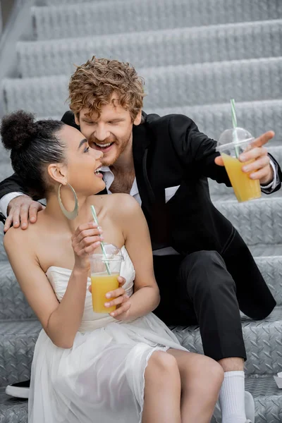 Cheerful redhead groom pointing with finger near african american bride with orange juice on street — Stock Photo