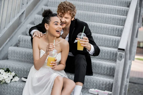 Sposa afroamericana eccitata con succo d'arancia che ride vicino allo sposo sulle scale all'aperto — Foto stock