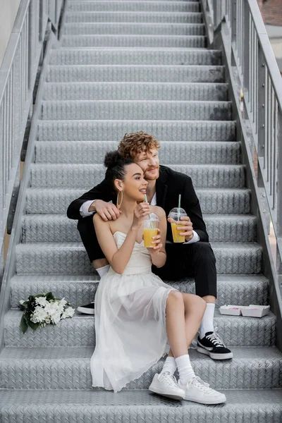 Happy interracial newlyweds with orange juice looking away on street stairs near bouquet — Stock Photo