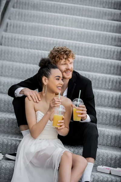 Redhead groom embracing happy african american bride sitting with orange juice on stairs in city — Stock Photo