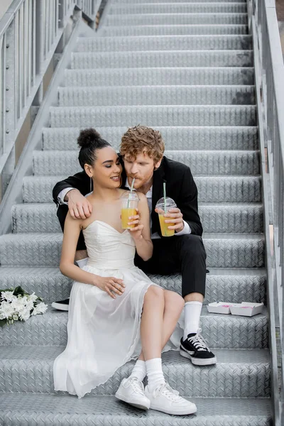 Redhead man drinking orange juice from straw near african american bride, sitting on stairs in city — Stock Photo