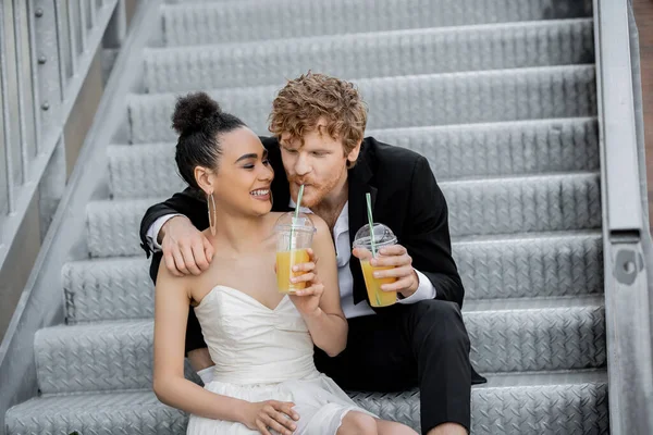 African american bride smiling near redhead groom drinking orange juice on stairs in city — Stock Photo