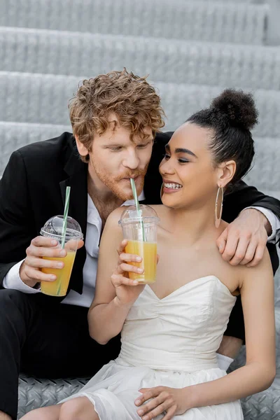 Pelirroja novio beber jugo de naranja de paja cerca alegre americano novia, boda en la ciudad, diversión - foto de stock