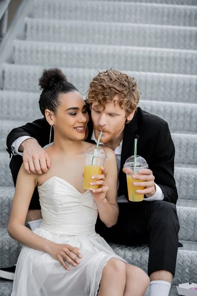 Mariage en ville, mariée afro-américaine souriant près du marié roux boire du jus d'orange de paille — Photo de stock