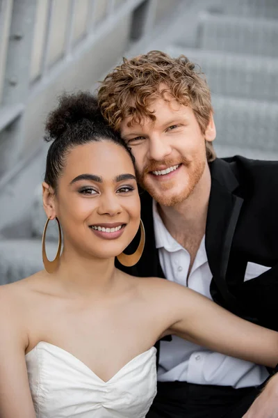 Retrato de casal multiétnico satisfeito e elegante de recém-casados olhando para a câmera ao ar livre — Fotografia de Stock