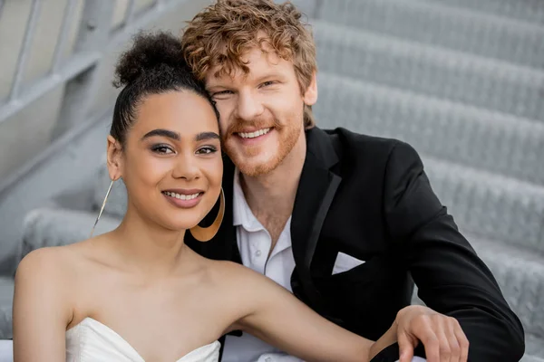Portrait de jeunes mariés interracial insouciants et élégants souriant à la caméra, mariage en plein air — Photo de stock