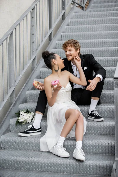 Interracial couple with sweet donuts sitting looking at each other on city stairs — Stock Photo