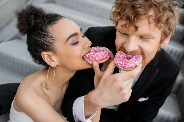 Young multiethnic newlyweds having fun and feeding each other with sweet donuts on urban street — Stock Photo