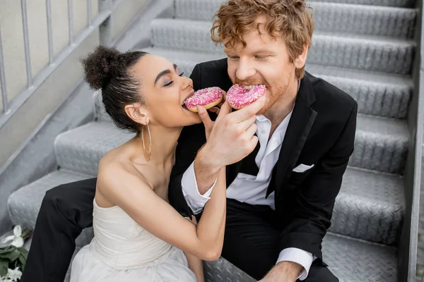 Celebración de la boda en la ciudad, pareja interracial alimentándose mutuamente con rosquillas dulces en las escaleras - foto de stock