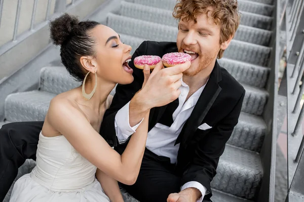 Romance urbaine, jeunes mariés multiethniques assis sur les escaliers et se nourrissant mutuellement de beignets sucrés — Photo de stock