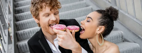 Mariage, cadre urbain, plaisir, couple de jeunes mariés interracial se nourrissant mutuellement avec des beignets, bannière — Stock Photo
