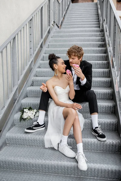 Hochzeit auf der Stadtstraße, fröhliches gemischtrassiges Paar sitzt auf der Treppe und isst süße Donuts — Stockfoto