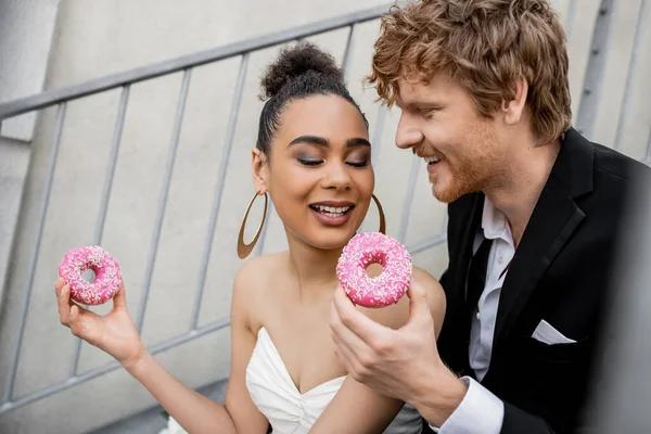 Delighted multiethnic couple in wedding outfit holding sweet donuts, wedding in urban setting — Stock Photo