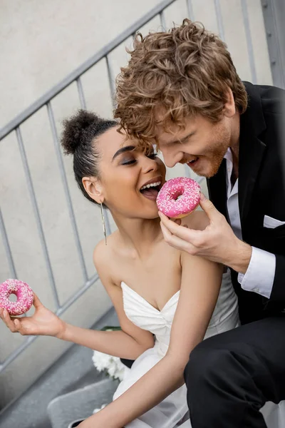 Веселий багатоетнічний молодята кусають солодкий пончик, весілля на міській вулиці — стокове фото