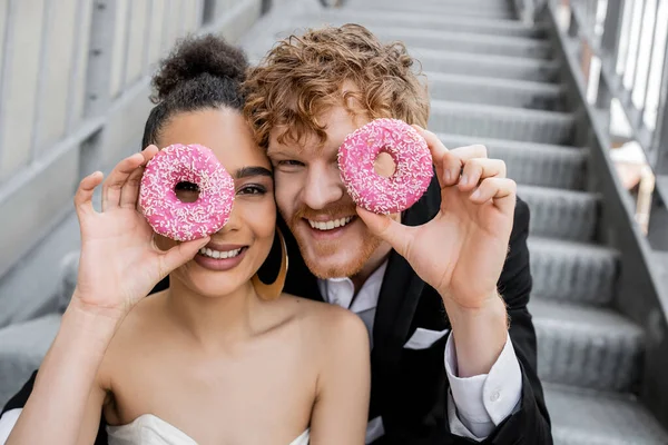 Having fun, wedding in city, excited interracial newlyweds obscuring face with donuts — Stock Photo