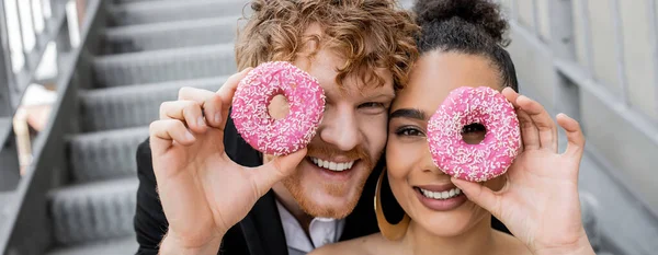 Outdoor wedding, happy interracial couple obscuring face with sweet donuts, banner — Stock Photo